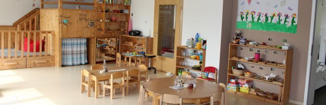 Zu sehen ist ein Gruppenraum mit zwei Tischen aus Holz für Kinder, mehreren Regalen mit Spielmaterial und einer Hochebene.