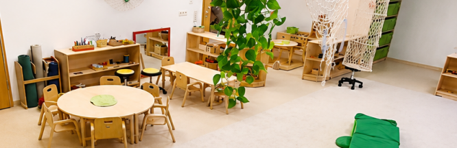 Spielraum für Kleinstkinder mit Pflanzen, Tischen, Stühlen, einem großen Spielteppich und mehreren Regalen mit verschiedenen Materialien.