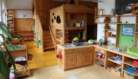 Zu sehen ist ein Gruppenraum. Auf der Ebene, von der aus das Bild gemacht wurde, sieht man eine Kinderküche und verschiedene Materialregale aus Holz. Im Hintergrund befindet sich eine hölzerne Spiel- und Hochebene.