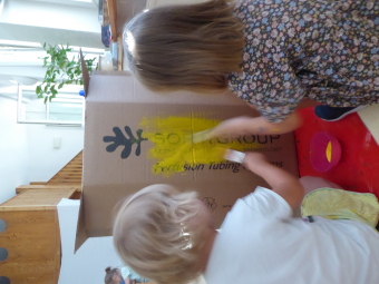 Zwei Kinder sitzen vor einem großen leeren Karton und malen diesem mit einem Pinsel gelb an.