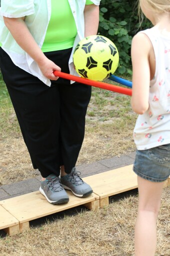 Eine erwachsene Person und ein Kind halten zusammen einen roten und einen blauen Stab in beiden Händen. Auf den Stäben balanciert ein gelb-schwarzer Ball. 