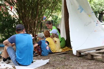 In einem Garten steht ein Zelt. Daneben befinden sich 2 erwachsenen Personen und drei Kinder und musizieren mit einer Gitarre. 