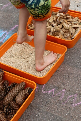 Orangefarbene Wannen stehen auf dem Boden und sind gefüllt mit Naturmaterialien. Kinderfüße ertasten das Material. 