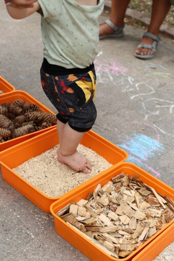 Orangefarbene kleine Wannen stehen auf dem Gartenboden und sind mit verschiedenen Naturmaterialien gefüllt. Kinderfüße ertasten das Material. 