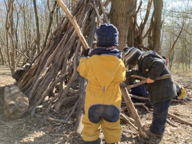 2 Kinder stehen mit dem Rücken im Bild. Vor ihnen sind lange Äste an einem Baum zu einem Zelt gestapelt