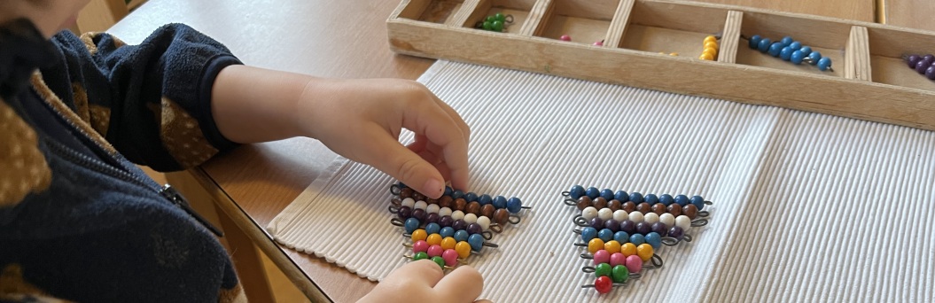 Zu sehen sind Kinderhände welche über einer Montessoriübung namens "bunte Perlenstäbschen" gehalten werden.