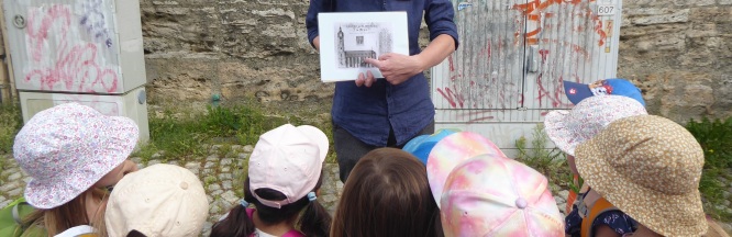 Kinder sind von hinten zu sehen, welche auf eine Fotografie in den Händen eines Museumsführers schauen. 