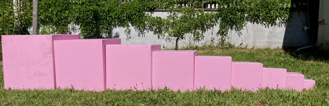 10 in ihrer größe abgestufte rosa Kuben liegen nebeneinander auf der Wiese. Von Groß zu Klein geordnet.