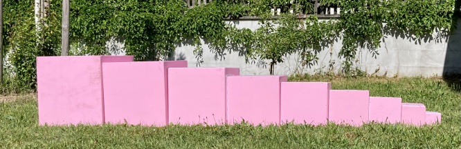 10 große rosafarbene Würfel liegen vom größten zum kleinsten Würfel auf einer Wiese aneinandergereiht. 