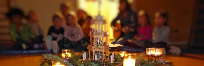 Eine Weihnachtspyramide aus Holz mit brennenden Kerzen steht im Vordergrund. Dahinter sind die Silhouetten singender Kinder zu sehen.