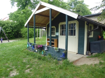 ein dunkelgrün gestrichenes Werkstatthaus mit einer kleinen Terasse und Geländer davor steht auf der Wiese im Garten. 