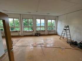 Ein leerer Kindergarten gruppenraum mit Blick auf die großen Fenster. auf dem Boden liegt Baustaub und eine Leiter steht im Zimmer. 