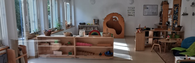 Zu sehen ist ein heller Gruppenraum mit kleinkindgerechten Material-Regalen und einer Kuschelhöhle aus Holz. 