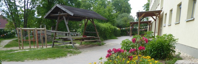 Am Hauseingang blühen bunte Blumen; links steht eine überdachte Sitzgruppe