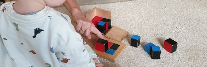 Auf einem weißen Teppich liegen rot-schwarz-blaue Holzbausteine, die zusammengesetzt einen Würfel ergeben. Im Bild setzen eine Pädagogin und ein Kind die Teile zusammen